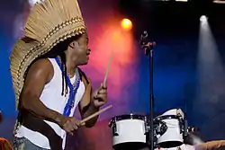 Carlinhos Brown stands on the left side of the image wearing a white shirt and a headpiece, holding drum sticks in both hands.  Two white drums are on the right side of the image with a microphone stand in between.