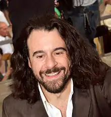 head and shoulders view of a smiling man with a dark beard and long hair, in a dark jacket with open white collar