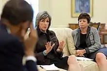 A pale-skinned woman in her fifties is sitting on a beige sofa in a large room with a window, a painting, and a bureau visible in the background. She has straight, tan-brown hair over her ears and down to her shoulder and is wearing a brown suit with a pale blue bracelet. She is gesturing with her hands and has a pen and a tablet of paper next to her. To one side of her is a light-brown-skinned woman in her fifties with brown hair, wearing a light-and-dark-grey suit and looking at the first woman. She is holding a pen and a writing pad is next to her as well. On the other side of the first woman is the out-of-focus image of the back of the head and shoulders and arm of a brown-skinned man with dark hair. He is looking at the first woman and holding his right hand to his temple, as if in pain or concentration.