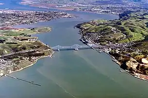 Rolling hills covered partially with buildings and roads frame the banks of a large channel of opaque water, which connects two larger bodies of water at the top left and bottom.