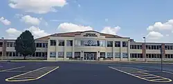 A photo of the front of a two story building with a creme colored facade and red brick columns every few feet.  The building has lots of windows.  Blue and grey letters in some of the windows spell the words "Cascade Cadets".  Benches and garbage cans flank the doors.  A large tree is visible to the viewer's left.