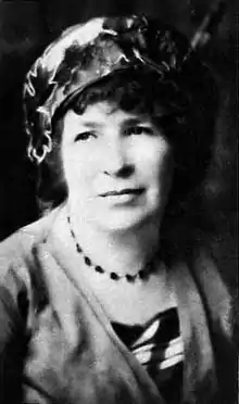 Portrait photo of Catherine Amy Dawson Scott. She is pale with dark darly hair that falls to her chin, a slight smile on her face as she gazes off toward the left, and is wearing an ornate flower hat, beaded necklace, and possibly a cardigan over a dress.