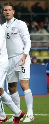 A dark-haired white man, wearing an all white football kit with number 5 imprinted on it.