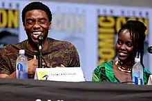 A man and woman sitting behind a panel desk, the man is laughing and the woman smiling.