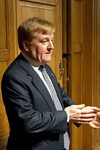 A suited middle-aged man with ginger hair standing in a room with wood paneling. He has a concerned expression and is rubbing his hands.