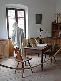 Photograph of a monk standing in a cell with a table, chair and books.