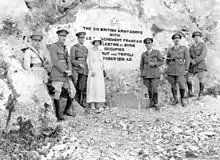 Five soldiers in Sam Browne belts, riding boots and peaked caps, and one woman in light coloured dress and matching hat in front of a stone inscription that reads: "The British XXI Corps with Le Detachment Français de Palestine et Syrie occupied Beirut and Tripoli October 1918 AD."