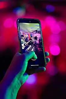 A hand holding a phone in which you can see a band performing live music.