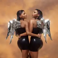 Chloe and Halle with shiny, metallic wings on their backs, both wearing latex dresses, standing and holding each other by one arm in front of a dusty background