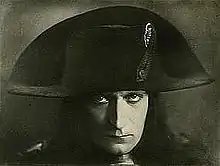 A monochrome photographic portrait of a handsome man in his late 20s wearing a French general's uniform from the 1790s and a cocked hat over stringy dark hair that reaches his shoulders