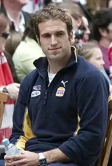 A man with brown hair in a navy blue jacket sits with his hands on his lap