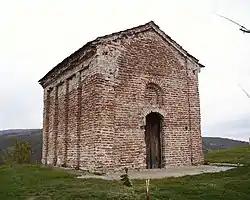 (Top image): A worn red brick building with decorative inlets on the side; the roof of the building is a gable roof; the door is a highly weathered wooden door with a decorative indent just above that is is the shape of a window. (Church of Saint John Orljane) (Bottom Image): Map of Serbia with the location of the settlement, in south eastern Serbia, marked by a red dot.