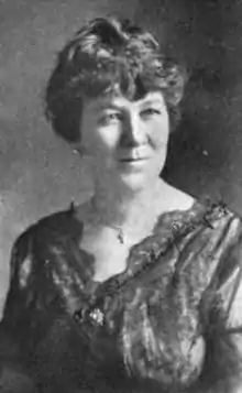 A middle-aged white woman, dark hair dressed up from her neck and shoulders, wearing a dark lace-trimmed dress with a v-neckline