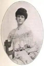 Young white woman, seated, wearing a light-colored lacy dress with a high collar, hair in an updo.