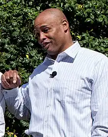 A man wearing a buttoned shirt stands in front of a vine and smiles.