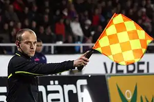 A man dressed in black. He is stretching his right arm forwards, holding a red and yellow chequered flag.