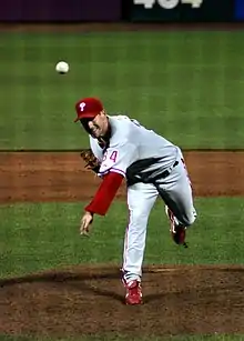 A left-handed man in a gray baseball uniform with red trim and a red baseball cap stands on a pitchers' mound having thrown a baseball toward the camera.