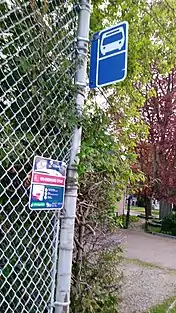 a bus stop sign for Cobourg Transit, attached to a pole holding up a fence of a elementary school playground. On the fence next to it is another sign advertising Cobourg Ride, an on demand transit pilot. There are plenty of differently coloured trees in the background of the image.