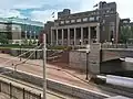 Washington Avenue's light rail tracks, pavement, and bus stop, in front of a retaining wall, on top of which sits Coffman's front lawn, with Coffman itself directly behind it. Slightly to the left of Coffman is the glass facade of Nils Hasselmo Hall.