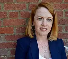 A woman with red hair and freckles standing against a red brick wall