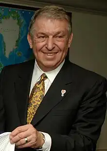 A man, wearing a black coat, white shirt and a tie, is posing for a photograph.