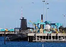 A submarine next to a dock, which is occupied by several cranes and other mechanical equipment