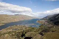 A big river and a nearby highway wind through a gorge between parallel ridges. The hills on the left are largely brown and treeless, while the hills on the right are partly covered by evergreen trees and grasses. A small lake lies in the foreground between the highway and the river.