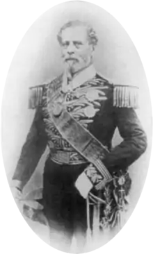 Tintype photograph depicting a full-length portrait of an older, bearded man wearing an elaborately embroidered military dress uniform with heavy epaulets, sash of office, sword, gloves and holding a bicorn hat in his right hand