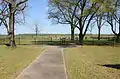 A small fenced cemetery with large trees on a flat, green landscape