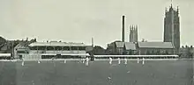 Black and white photograph of a cricket ground with players on the field dressed in white. In the background two churches, a tall industrial chimney and some houses can be seen.