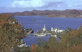 A small harbour surrounded by trees offers shelter to several yachts, fishing boats and a motor launch. The harbour is connected to the sea beyond by a lock gate. A small skerry lies in the water and brown rocky hills dominate the distance.