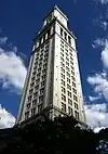 Angled, ground-level view of white limestone tower. Near the building's pyramidal-cap, a large, 4-faced clock is visible