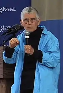 Image of Cynthia Enloe standing with a mic in her hand wearing a blue coat.