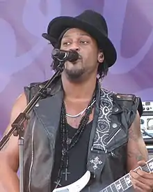 An African-American man singing into a microphone. He is wearing a black hat and a sleeveless leather jacket.