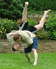 Two men in combat appear on a green lawn in front of a shrubbery. Both men are wearing dark coloured shorts and cream coloured tops. One man has a tactical advantage, and is throwing his opponent head-first towards the ground.