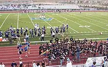 A high school football field with a large "DV" emblem in the center of the turf. Dougherty Valley football players in blue and silver uniforms are lined up on the side of the field. On the track surrounding the field, there are cheerleaders in light blue, white, and navy blue uniforms with blue pom-poms to the left. To the right of the track there are members of the pep band with their instruments in black shirts.