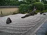 Rock garden with raked gravel and large standing stones in front of a small wall and green plants.