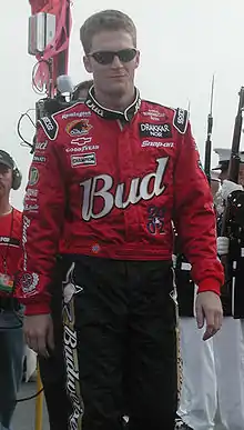 A man in his late twenties wearing black sunglasses and a red and black racing suit adorned with sponsors' logos