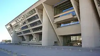 A tall beige building with an angled front face, leaning out from the top, is supported by three columns and covered with rows of windows