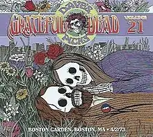 Two skeletons, a man and a woman, lying together in the grass and flowers, with the Boston skyline in the background