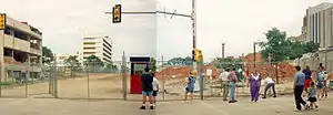 Two images are stitched together showing the site of where the building stood prior to its demolition. A crowd of people are visible in front of the chain link fence blocking entrance to the site. Large piles of dirt can be seen on the site as well as damage to nearby buildings.