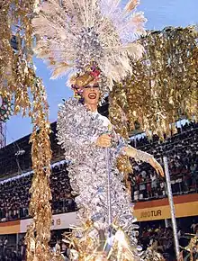 Rogéria standing on top of a platform with a dress covered in reflective silver feathers and a hat with peacock feathers.