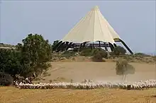 A large sand-coloured conical tent on a small hill before a pale blue sky, with a few trees and a herd of sheep on dry grassland in the foreground