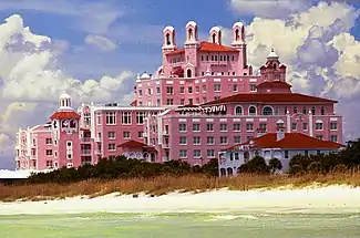 A pink, multi-floor hotel stands out above a calm sea and a windswept beach.