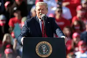 Donald Trump speaking at a podium. Behind him is a crowd.