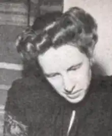 A white woman with hair in a bouffant updo, looking downward, wearing a dark blouse or dress