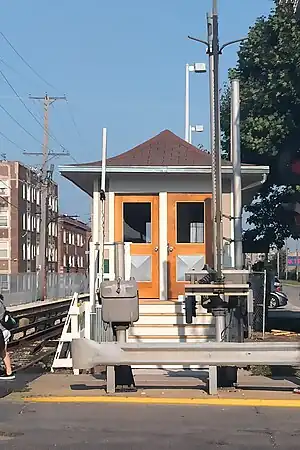 A beige Craftsman station house, situated at the right of two tracks, with a red peaked roof and a double beige door.