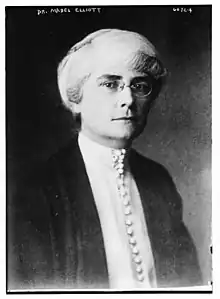 A white woman with white hair and dark eyebrows, wearing eyeglasses, a high-collared white blouse with many fabric-covered buttons down the front, and a dark jacket