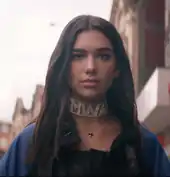 Dua Lipa walking on the street wearing a "Mwah" choker and a blue jacket.