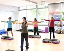 Four people exercise with dumbbells in the hands, standing behind plastic step tools.